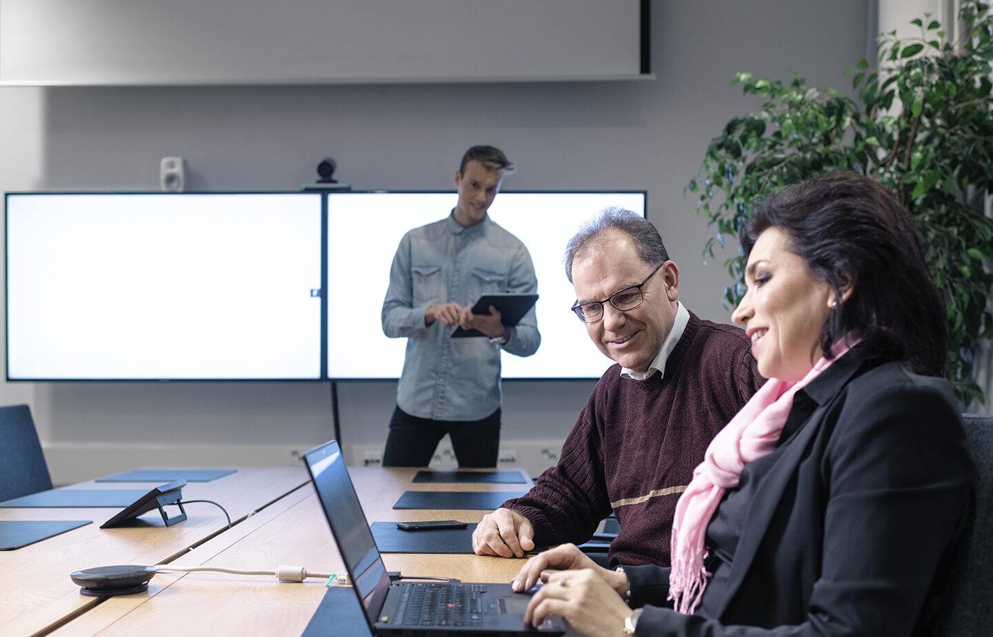 Kaksi henkilöä kuuntelemassa yhtä presentoijaa EPV:n neuvotteluhuoneessa. Kuuntelijat katsovat samalla neuvottelupöydällä olevan kannettavan tietokoneen ruutua. Presentoijalla on kädessään taulutietokone ja takanaan kaksi suurta näyttöä.