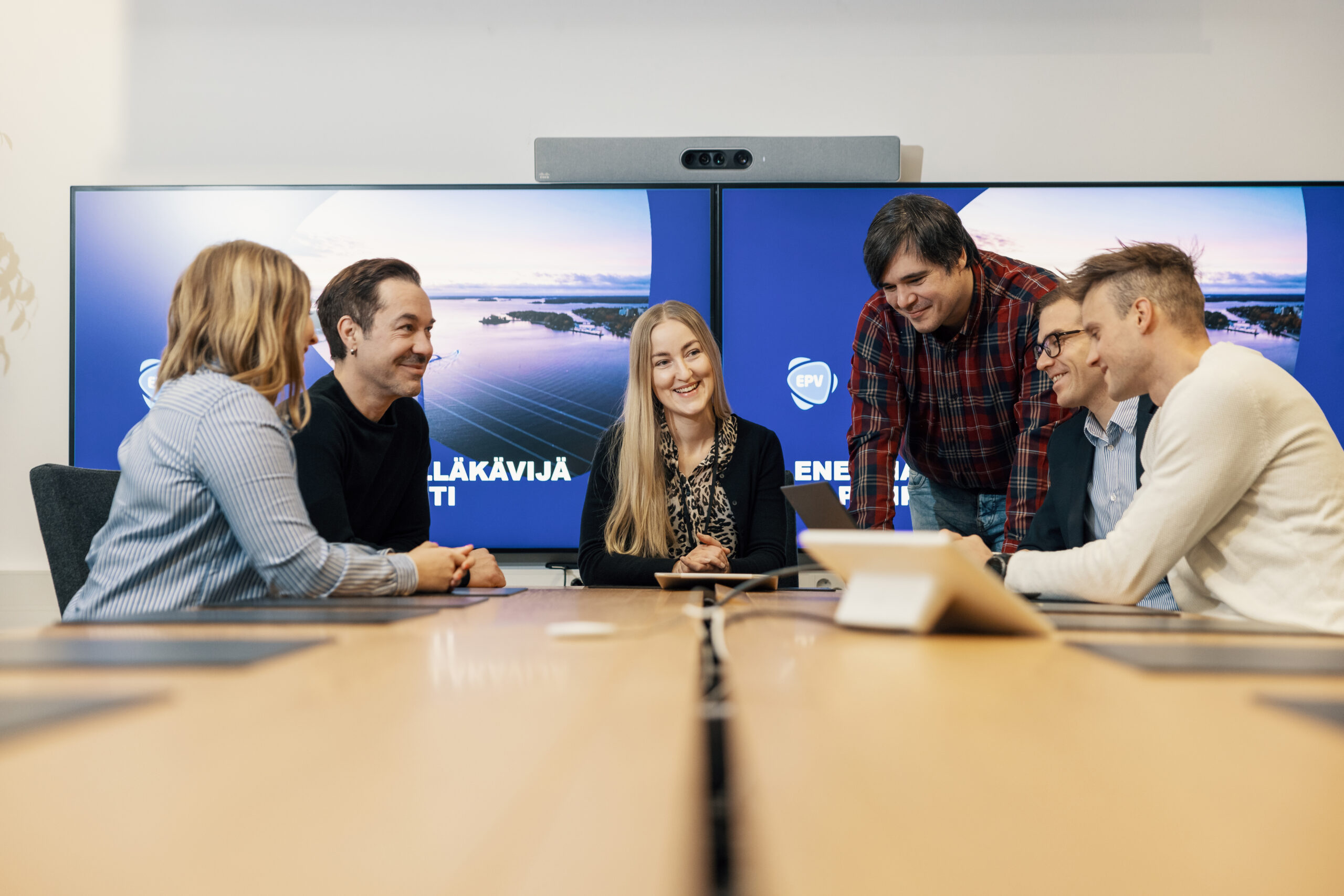 Kuusi henkilöä katsomassa taulutietokoneen ruutua EPV:n neuvotteluhuoneen pöydän äärellä. Pöydällä on myös kannettava tietokone. Henkilöiden takana on kaksi suurta näyttöä, joissa molemmissa lukee ”energia-alan edelläkävijä ja pelikentän tähti”.
