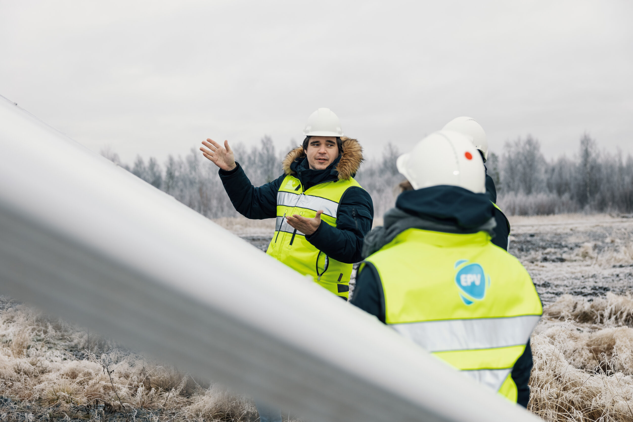 Kolme henkilöä juttelemassa suuren aurinkopaneelin edessä huomioliivit päällään ja suojakypärät päässään.