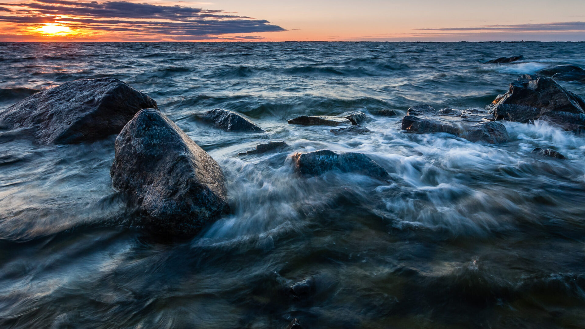Merinäkymä upeassa auringonlaskussa. Etualalla kiviä, joihin aallot lyövät.